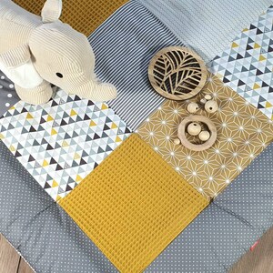 Può includere: Un tappetino da gioco per bambini grigio e giallo con una varietà di motivi geometrici. Il tappetino presenta un giocattolo di peluche a forma di elefante, una decorazione in legno a forma di foglia e una ciotola di legno con perline di legno.