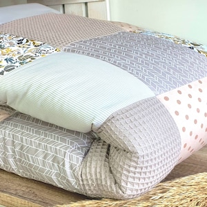 Puede incluir: Un edredón de patchwork con varios patrones y colores de tela. El edredón presenta secciones de estampado floral, tela de rayas, patrones de puntos y diseños geométricos en tonos de blanco, gris y rosa. El edredón está doblado y parece estar sobre una estantería de madera.