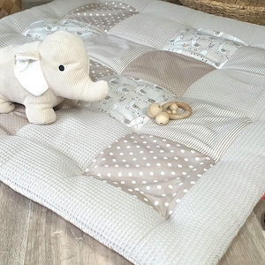 Może przedstawiać: Mata do zabawy w patchworku w kolorze beżowym i białym z pluszową zabawką w kształcie słonia i drewnianymi gryzakami. Mata wykonana jest z miękkiej tkaniny i ma pikowaną fakturę.