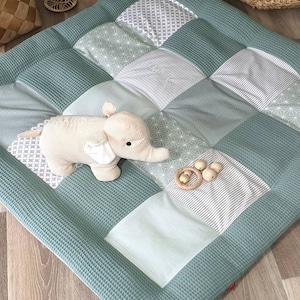Może przedstawiać: Zielona i szara mata do zabawy w patchwork z pluszową zabawką w kształcie słonia i drewnianymi gryzakami.