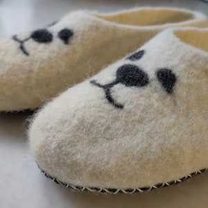 May include: A pair of white wool slippers with black felt animal faces. The slippers have a black felt trim around the edges.