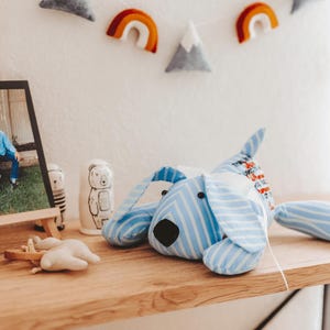 May include: A blue and white striped dog-shaped plush toy with floppy ears and a black nose rests on a wooden shelf. The toy is part of a nursery decor set, alongside a framed photo, wooden airplane, and decorative bear figurines.