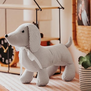 May include: A stuffed dog toy made of fabric with a white and black grid pattern. The dog has floppy ears, a small tail, and a white ribbon tied around its neck. The toy is standing on a striped surface.