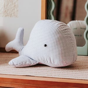 May include: A stuffed whale toy made of white fabric with a black grid pattern. The whale has a black button eye and a tail and flipper fins. The toy is resting on a striped surface.