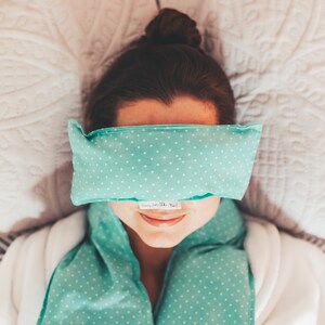 May include: A person wearing a teal blue eye mask with white polka dots and a matching neck wrap. The eye mask has the text "Sleepy from the Head" printed on it.