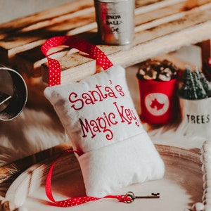May include: A white fabric Christmas stocking with red embroidered text reading "Santa's Magic Key." A small metal key is attached to a red polka dot ribbon. Other Christmas decorations include a miniature bucket with "Snow Balls 5¢" text.