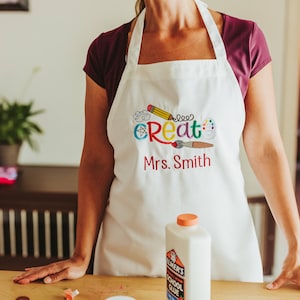 May include: White apron with the word "create" in colorful letters and a pencil, paintbrush, and paint palette design. The apron also has the name "Mrs. Smith" embroidered on it.