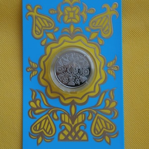 May include: A silver coin with a floral design is displayed in a blue and yellow packaging with the text "National Bank of Ukraine" and "örnek П орynek" printed on it.