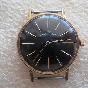 Puede incluir: Un reloj de pulsera vintage en tono dorado con esfera negra. El reloj presenta manecillas e indicadores de hora dorados. La esfera del reloj tiene el texto "ЛУЧ" y "23 камня". El reloj tiene una corona dorada y una correa de acero inoxidable.