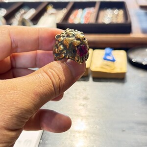 K&ouml;nnte beinhalten: Ein goldfarbener Ring mit einem detaillierten L&ouml;wenkopf-Design und einem roten Edelstein im Maul. Der Ring wird pr&auml;sentiert, um die filigrane Handwerkskunst und die leuchtende Farbe des Edelsteins zu zeigen.