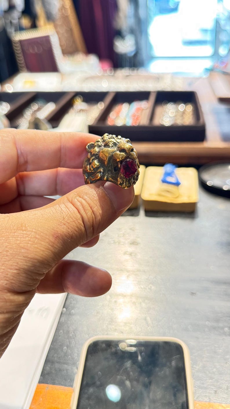 K&ouml;nnte beinhalten: Ein goldfarbener Ring mit einem detaillierten L&ouml;wenkopf-Design und einem roten Edelstein im Maul. Der Ring wird pr&auml;sentiert, um die filigrane Handwerkskunst und die leuchtende Farbe des Edelsteins zu zeigen.