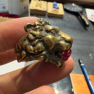 Può includere: Un anello dettagliato in stile antico con la testa di un leone e una pietra color rubino in bocca. L'anello è in metallo dorato. Un gioiello unico, tenuto in mano da una persona.
