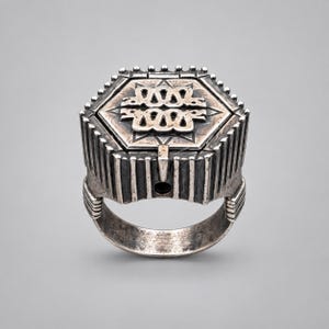 May include: A silver ring featuring a large hexagonal top with a raised, intricate, symmetrical pattern of interlocking loops and star-like points. The sides of the ring are decorated with vertical grooves, and the band has a weathered, oxidised finish.
