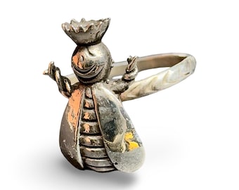 May include: A silver-colored bee-shaped ring. The bee has a smiling face, a crown, and outstretched arms. The ring band is a simple, twisted design. The bee's wings and body have detailed textures.