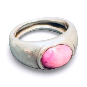 Könnte beinhalten: Ein silberfarbener Ring mit einem abgerundeten Band und einem großen, ovalen, rosa Stein. Der Ring hat eine glatte, polierte Oberfläche und ein klassisches Design, geeignet für den Alltag oder besondere Anlässe.
