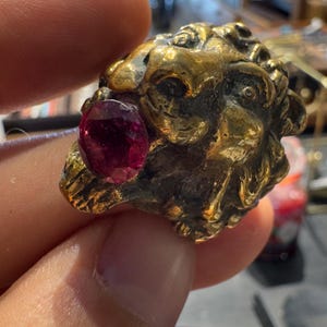 K&ouml;nnte beinhalten: Ein goldfarbener Ring mit einem detaillierten L&ouml;wenkopf-Design und einem prominenten, oval geformten, tiefroten Edelstein. Die filigranen Details des Rings und der leuchtende Edelstein bilden einen auff&auml;lligen Kontrast, der die Handwerkskunst hervorhebt.