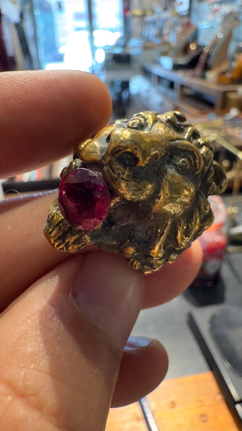 K&ouml;nnte beinhalten: Ein goldfarbener Ring mit einem detaillierten L&ouml;wenkopf-Design und einem prominenten, oval geformten, tiefroten Edelstein. Die filigranen Details des Rings und der leuchtende Edelstein bilden einen auff&auml;lligen Kontrast, der die Handwerkskunst hervorhebt.