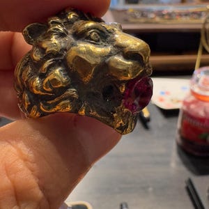 K&ouml;nnte beinhalten: Ein goldfarbener L&ouml;wenkopfring mit einem rubinroten Edelstein im Maul. Der Ring ist mit strukturiertem Fell und Gesichtsz&uuml;gen versehen. Der Ring wird von den Fingern einer Person gehalten.