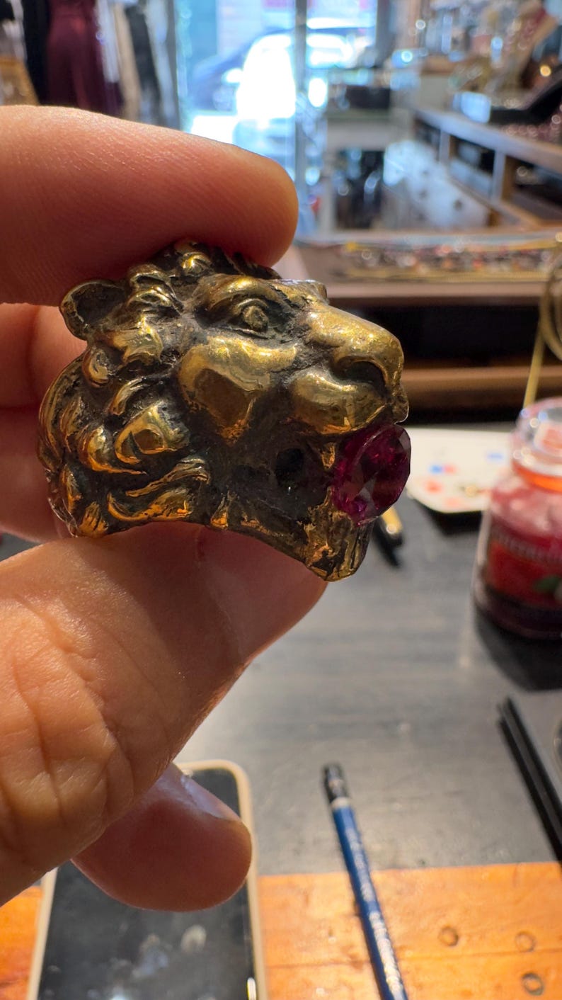 K&ouml;nnte beinhalten: Ein goldfarbener L&ouml;wenkopfring mit einem rubinroten Edelstein im Maul. Der Ring ist mit strukturiertem Fell und Gesichtsz&uuml;gen versehen. Der Ring wird von den Fingern einer Person gehalten.