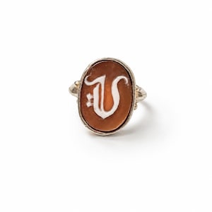 Op de afbeelding: Een zilveren ring met een ovale, roodbruine steeninleg. De steen is gegraveerd met een witte, gestileerde letter V in gotisch schrift. De metalen ring heeft een gepolijste afwerking met kleine decoratieve bolletjes bij de zetting.
