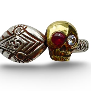 Può includere: Un anello in due parti con un teschio dorato con una gemma rossa e un occhio di gemma trasparente, collegato a un design geometrico argentato. L'anello ha una fascia attorcigliata.