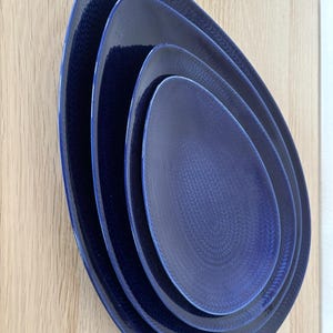 Puede incluir: Juego de cuatro platos de cerámica azul con un patrón tejido. Los platos son de forma ovalada y están anidados uno dentro del otro.