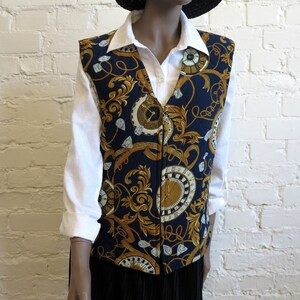 May include: A navy blue vest with a gold and white clock and scroll pattern. The vest is worn over a white collared shirt. The vest has a V-neck and a zipper closure. The vest is a vintage style.
