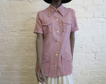 Può includere: Una camicia a maniche corte con un motivo a quadri rossi, bianchi e blu navy. La camicia ha un colletto, due tasche sul petto e due tasche inferiori. Presenta bottoni in legno ed è esposta su un manichino.
