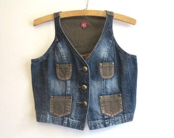 May include: A denim vest with a faded blue wash, featuring four pockets and brass-colored buttons. The vest has a brown interior lining and is hanging on a wooden hanger. The vest is a vintage style.