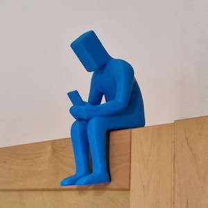 Könnte beinhalten: Eine blaue, 3D-gedruckte Figur eines sitzenden Menschen, der den Kopf gesenkt und auf ein Telefon schaut. Die Figur sitzt auf einer Holzoberfläche.