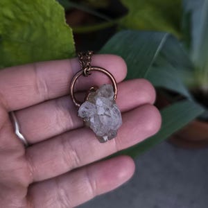Puede incluir: Un collar con colgante de color cobre con un grupo de cristales de cuarzo transparente. Los cristales están engarzados en un marco circular, suspendidos de una delicada cadena. El collar se sostiene en una mano, con follaje verde al fondo.