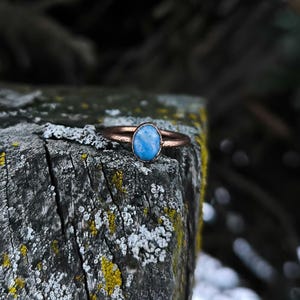 Könnte beinhalten: Ein kupferfarbener Ring mit einem ovalen, hellblauen Edelstein. Der Ring befindet sich auf einer verwitterten Holzoberfläche mit Flechten und Moos.