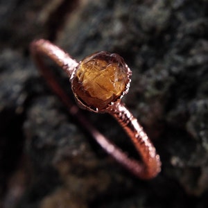 Op de afbeelding: Een ring van koperdraad met een ruwe, bruine en oranje edelsteen in het midden.