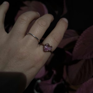 May include: A silver band ring and a copper ring with a pink gemstone are displayed on a hand. The gemstone has a rough, irregular shape. The background is dark with hints of pink.