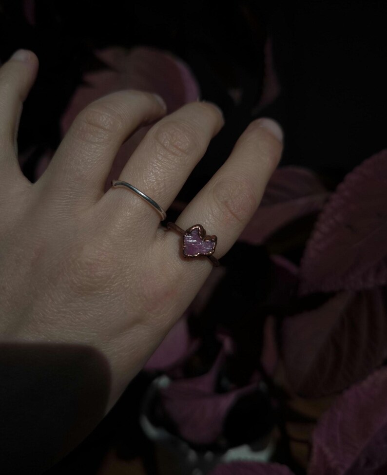 May include: A silver band ring and a copper ring with a pink gemstone are displayed on a hand. The gemstone has a rough, irregular shape. The background is dark with hints of pink.