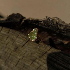 Könnte beinhalten: Ein Ring mit einem rohen, grünen Peridot-Edelstein und einem kupferfarbenen Band. Der Edelstein hat eine unregelmäßige Form und ist in eine kupferfarbene Lünette gefasst. Der Ring wird auf einem Stück verwittertem Holz präsentiert.