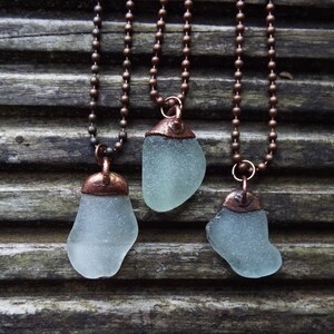 Puede incluir: Tres collares con cadenas de cobre y colgantes de vidrio de mar. El vidrio de mar es de color azul claro y tiene un acabado esmerilado. Las cadenas de cobre son de color marr&oacute;n oscuro.