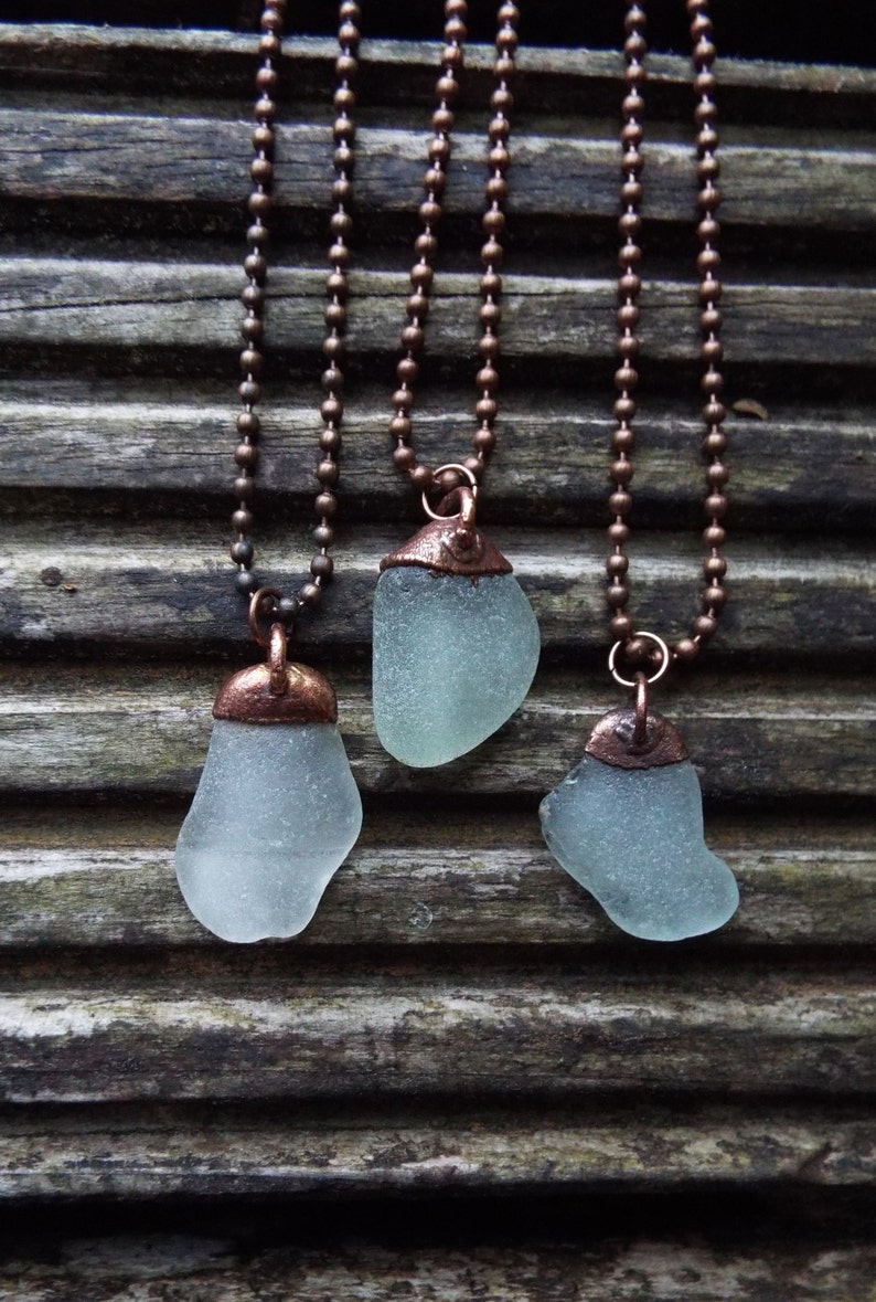 Puede incluir: Tres collares con cadenas de cobre y colgantes de vidrio de mar. El vidrio de mar es de color azul claro y tiene un acabado esmerilado. Las cadenas de cobre son de color marr&oacute;n oscuro.