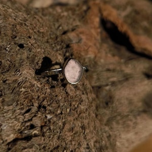 Könnte beinhalten: Ein silberner Ring mit einem hellrosa, oval geformten Stein. Der Ring ist auf einer strukturierten, braunen Holzoberfläche platziert, was einen Kontrast zwischen dem glatten Stein und dem rauen Holz erzeugt.