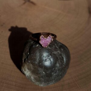 May include: A copper-colored ring with a heart-shaped, rough-cut pink gemstone. The ring sits atop a dark gray, rounded stone, set against a wooden background. The ring's design is simple, highlighting the unique gemstone.