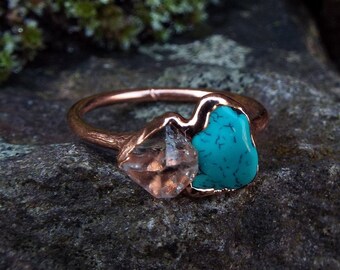 May include: A rose gold ring featuring a turquoise and clear quartz stone set in the band. The ring is displayed on a dark grey rock background.