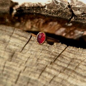 May include: A gold ring featuring an oval, faceted, red gemstone. The ring is set in a gold bezel and is placed on a piece of weathered wood. The gemstone has a deep red hue and reflects light.