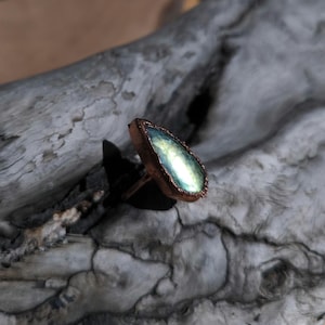 Könnte beinhalten: Ein kupferfarbener Ring mit einem birnenförmigen Labradoritstein, der schillernde Blau-, Grün- und Gelbtöne aufweist. Der Ring ist auf Treibholz platziert, wodurch die natürlichen Farben des Steins und die warmen Töne des Metalls hervorgehoben werden.