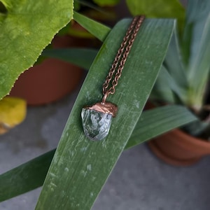 Puede incluir: Collar con un colgante de cristal verde en bruto, suspendido de una cadena color cobre. El cristal, de forma irregular, está engastado en un bisel de cobre. El collar descansa sobre una gran hoja verde.