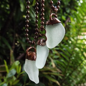 Puede incluir: Tres colgantes de vidrio de mar en cadenas de cobre. El vidrio de mar es de color blanco lechoso claro y las cadenas de cobre son de color marr&oacute;n c&aacute;lido.