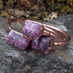 Może przedstawiać: Trzy miedziane pierścionki z surowymi fioletowymi kamieniami szlachetnymi osadzonymi w metalu. Pierścionki są ułożone jeden na drugim.