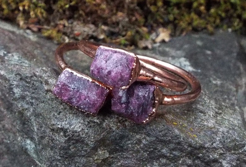 May include: Three copper rings with raw purple gemstones set in the metal. The rings are stacked on top of each other.