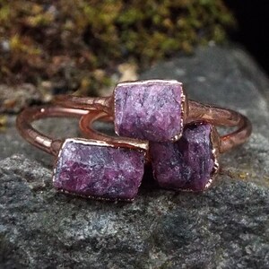 May include: Three copper rings with raw purple amethyst stones set in the metal. The rings are stacked on top of each other.