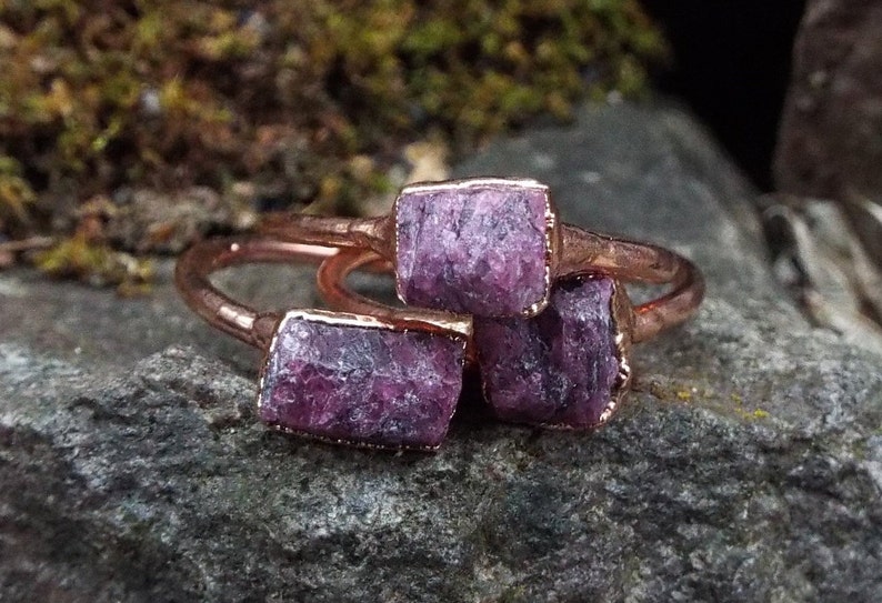 May include: Three copper rings with raw purple amethyst stones set in the metal. The rings are stacked on top of each other.