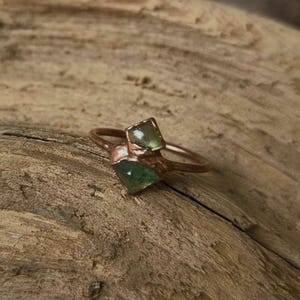 May include: A handcrafted copper ring featuring two square-cut, green gemstones. The ring's band and setting have a rustic, organic design, showcasing the natural beauty of the materials. The ring is displayed on a textured, wooden surface.